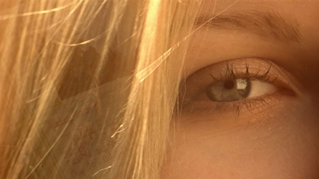 An extreme close-up of a teenage girl's green right eye, with blonde hair falling down next to it.